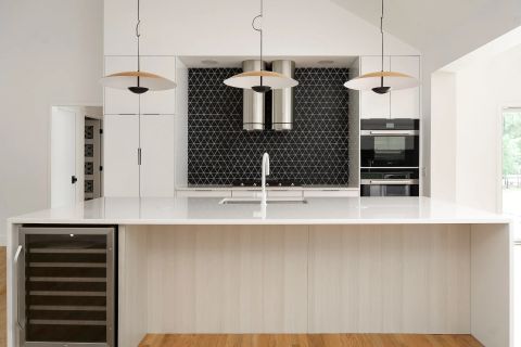 Aiken modern kitchen with white cabinetry, white quartz island with light wood base and wine cooler, black geometric tile backsplash, dual range hoods, and double ovens by Tekton Builds.