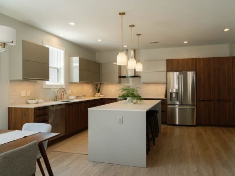 Greenville kitchen with gray island, walnut cabinetry, taupe upper cabinets, three gold-and-white pendant lights, black bar stools, and stainless steel appliances by Tekton Builds.