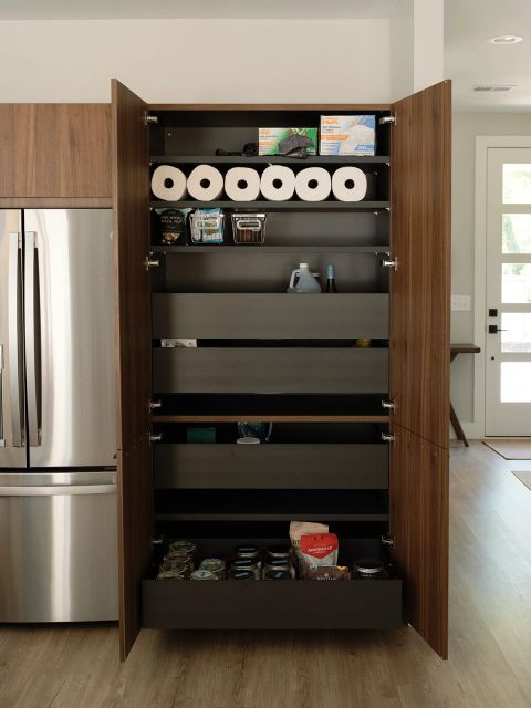Greenville kitchen pantry with open walnut doors, dark interior shelving, pull-out drawers with paper towels and dry goods beside stainless steel refrigerator by Tekton Builds.
