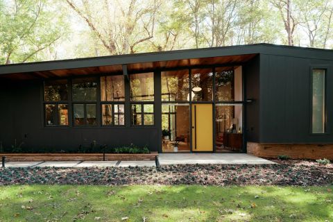 Greenville modern home with dark charcoal exterior, vibrant yellow front door, black-framed windows, exposed wood soffit, and wooded landscape by Tekton Builds.