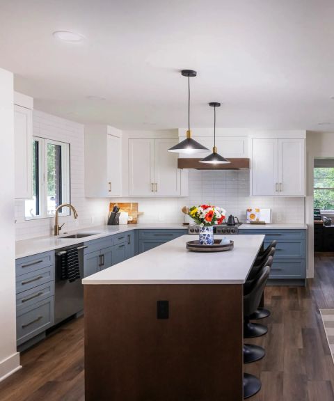 Remodeled kitchen with white upper cabinets, blue-gray lower cabinets, subway tile backsplash, wood-base island with bar stools, and brass pendant lights.